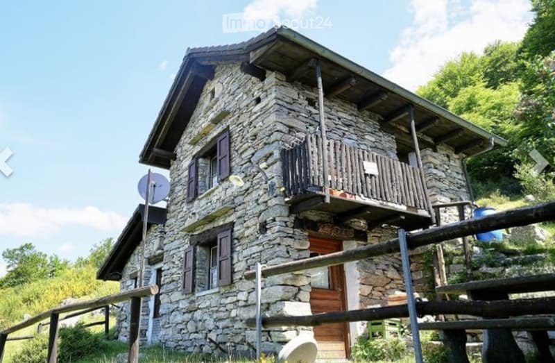 Two-story stone house with wooden balcony and shutters, surrounded by greenery.