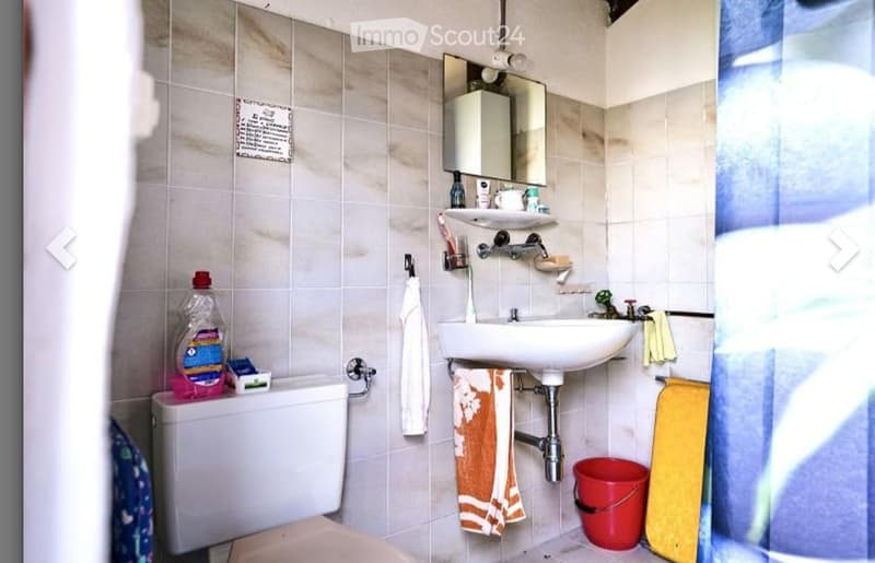 A small bathroom with a toilet, sink, mirror, and various personal care items. The walls are tiled, and towels are hanging on the sides.