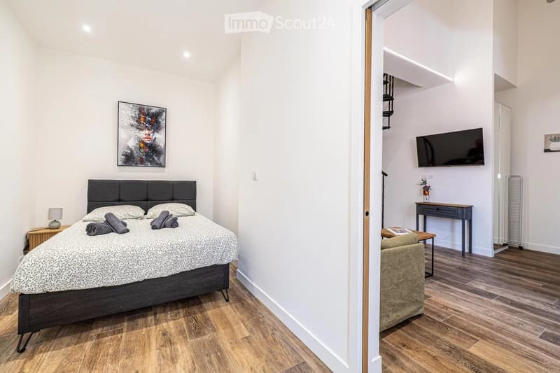 Cozy bedroom with a double bed, nightstand, lamp, and a small seating area with a chair and table. A flat-screen TV is mounted on the wall in the adjacent space.