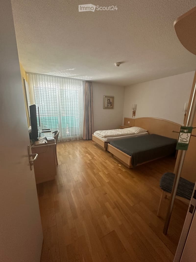 A furnished room with two single beds, a desk with a screen, a chair, and a picture on the wall. The room has wooden flooring and large windows with blinds.