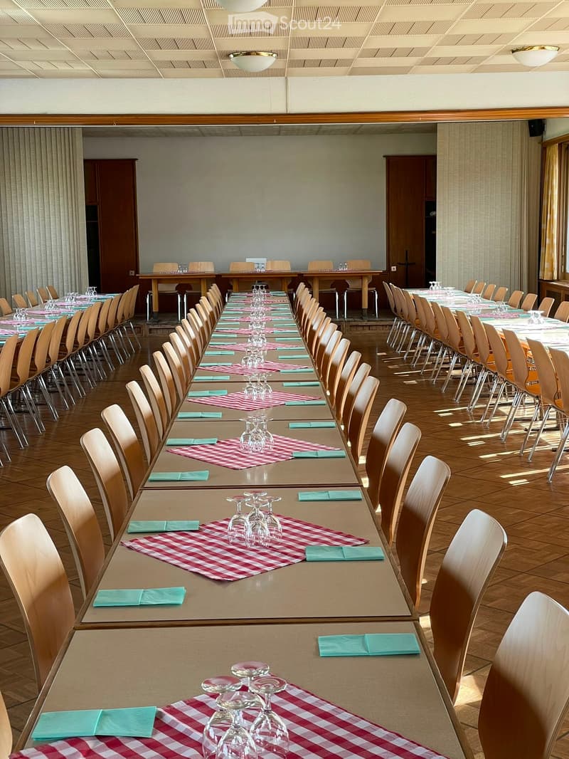 A large, well-lit dining hall with multiple long tables set for a formal event, each adorned with red and white checkered tablecloths, green napkins, and wine glasses.