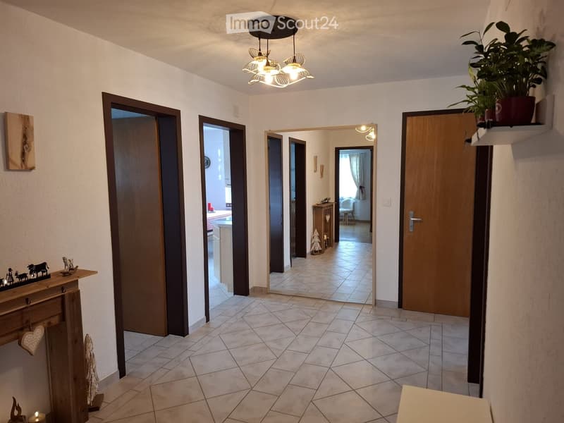 A well-lit hallway with tiled flooring, several wooden doors, a chandelier, and a small wooden console table with decorative items.