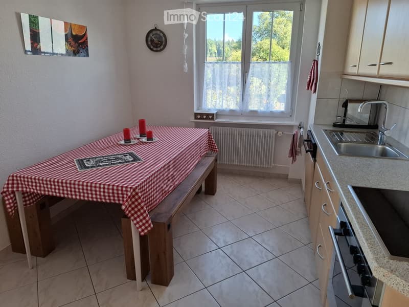 Bright kitchen with tiled floor, wooden table with red checkered tablecloth, sink, oven, cabinets, and a window with sheer curtains.