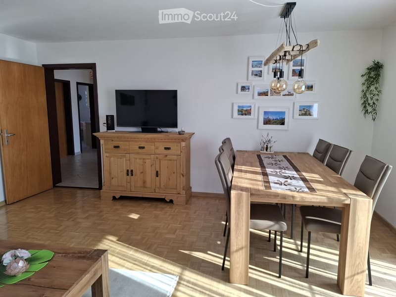 Bright dining room with wooden flooring, a large wooden dining table surrounded by six chairs, a wooden cabinet with a TV on top, and a decorative table runner. The room features framed pictures on the wall and a hanging light fixture.
