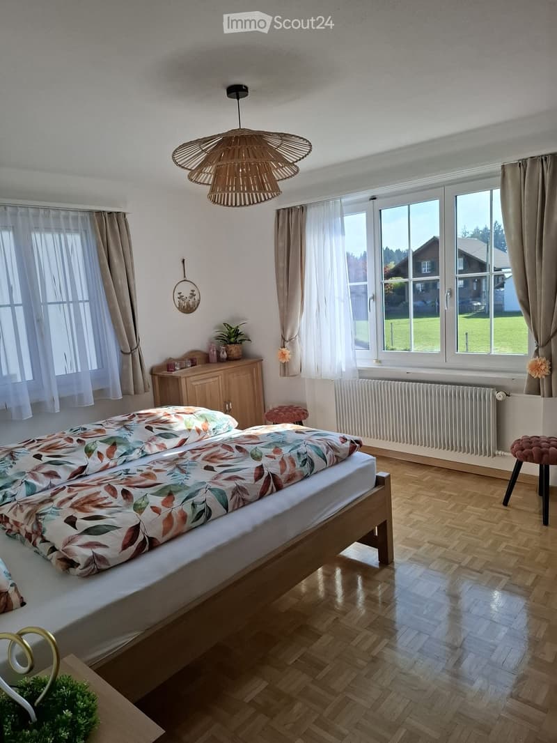 Bright bedroom with wooden flooring, large windows with curtains, a double bed with a leafy duvet, a nightstand with a plant, a wicker pendant light, and a radiator below the windows.
