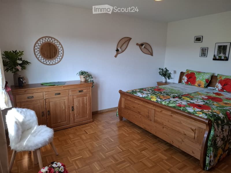 Cozy bedroom with wooden flooring, a large wooden bed with floral bedding, a wooden dresser, a white fur chair, and decorative wall mirrors.