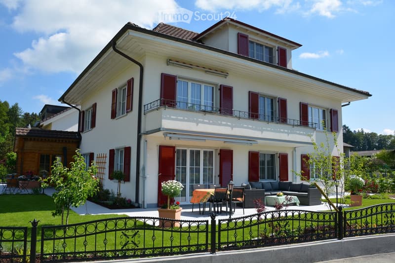 Two-story house with white exterior, red shutters, and a balcony on the upper floor. The front yard features a well-maintained lawn, a black wrought-iron fence, and outdoor seating including a sofa, coffee table, and dining table with chairs.