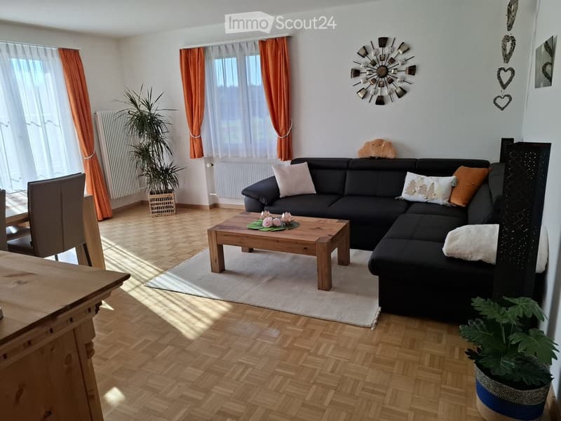 Bright living room with wooden herringbone flooring, large windows with orange curtains, black sectional sofa with multiple throw pillows, wooden coffee table with floral centerpiece, and potted plants.