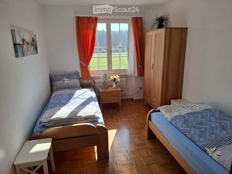 Cozy bedroom with two single beds, wooden nightstand, lamp, flower vase, wardrobe, and a small stool.