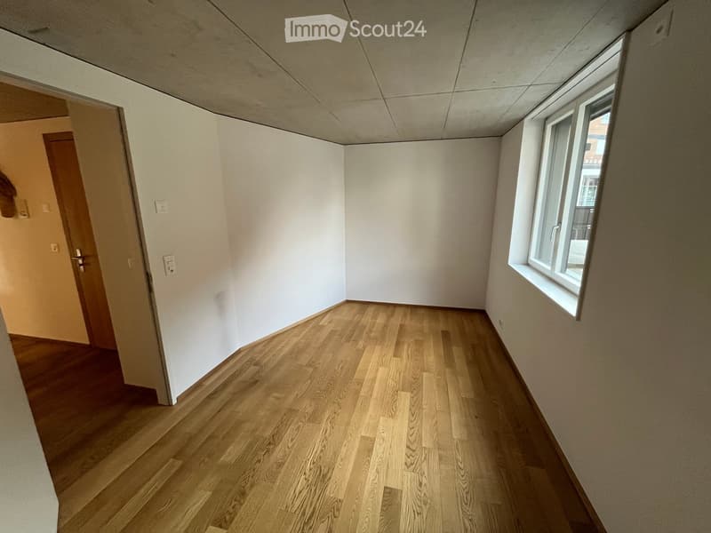 Empty room with light wooden flooring, white walls, and a single window on one side. The room has a door leading to another area.
