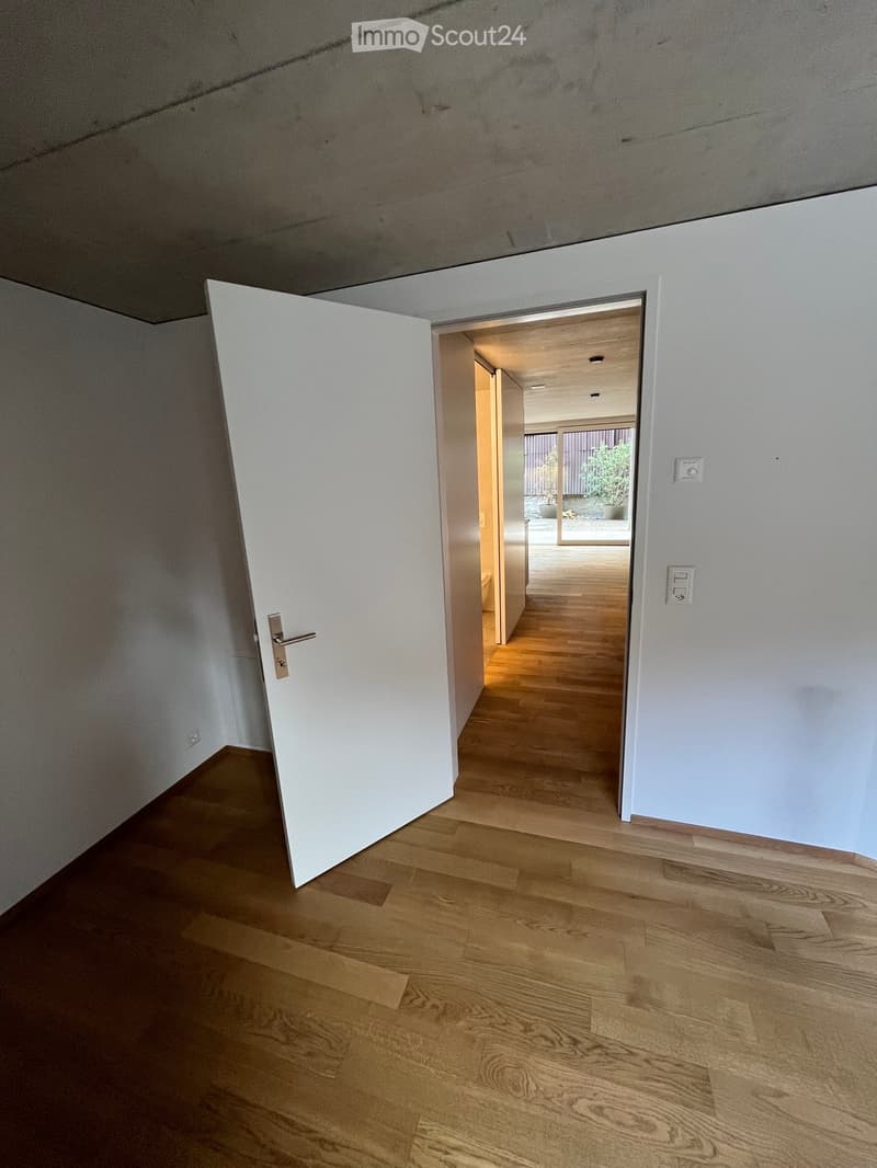 Empty hallway with light wooden flooring, white walls, and an open door leading to another room.