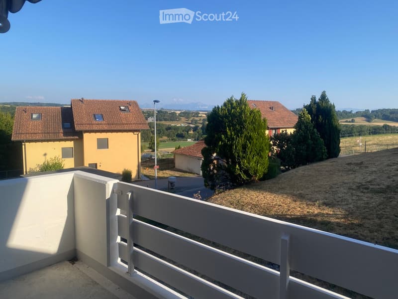 Balcony with white railing overlooking a rural landscape with houses, trees, and open fields.