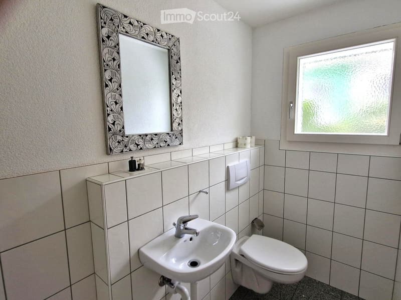 A small bathroom with white tiled walls and floor, featuring a sink, toilet, mirror with decorative frame, and a window with frosted glass.