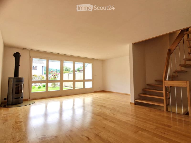 Bright, empty room with wooden flooring, large windows, and a staircase leading upwards. A wood-burning stove is visible on the left side.