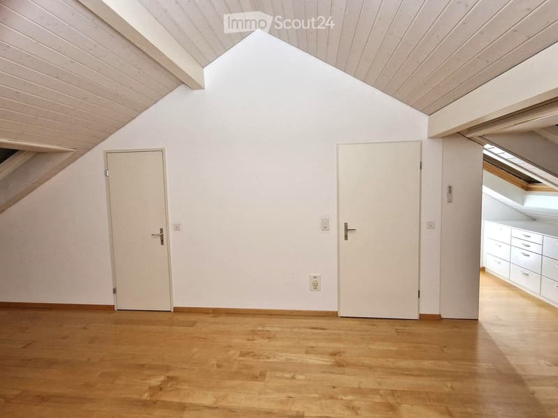 Empty room with wooden flooring, white walls, and two doors. The room has a slanted wooden ceiling with skylights, and a small section of a kitchen with white cabinets is visible through one of the doors.
