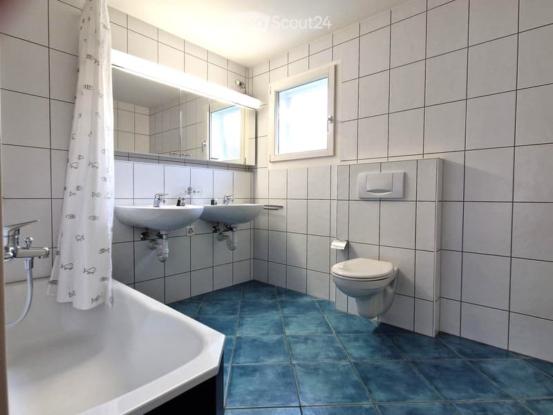 Modern bathroom with two sinks, a bathtub, a toilet, and a large mirror above the sinks. The walls are tiled in light grey, and the floor is covered in blue tiles.