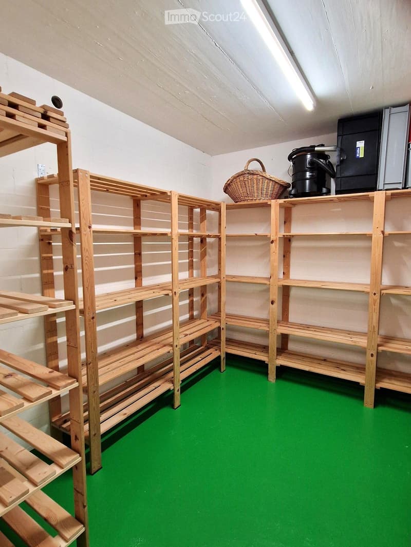 Empty storage room with green flooring, wooden shelving units, and a vacuum cleaner on top of a shelf.