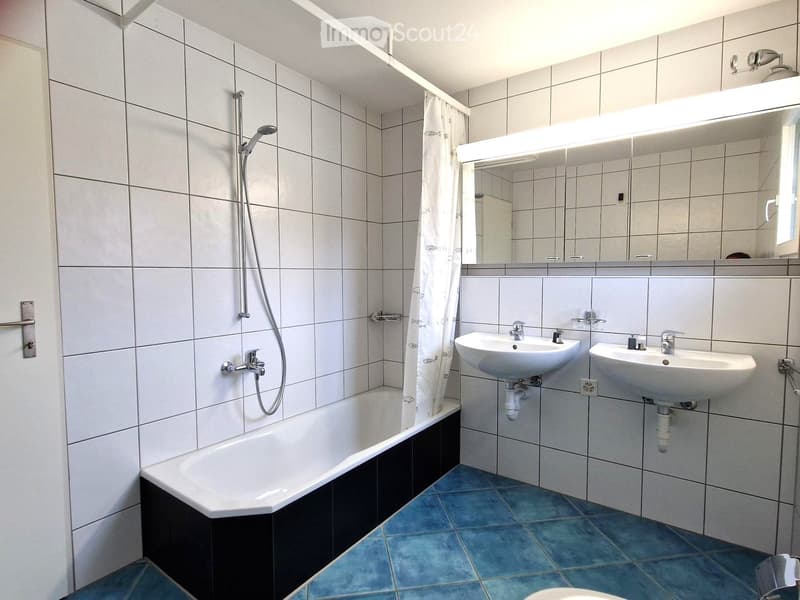 Modern bathroom with white tiled walls, blue tiled floor, bathtub, two sinks, large mirror, and shower area with a curtain.