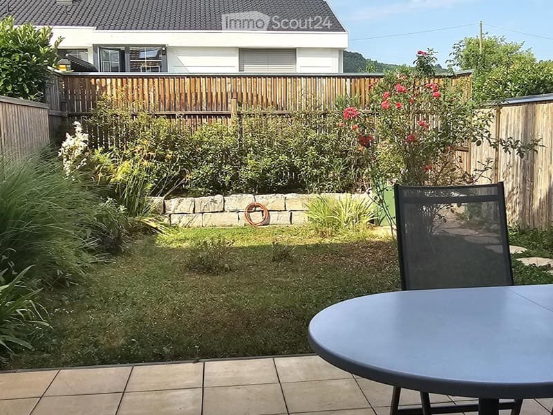 Well-maintained garden with a stone wall, wooden fence, and a patio area with a round table and chair. The background shows a modern house with large windows and a balcony.