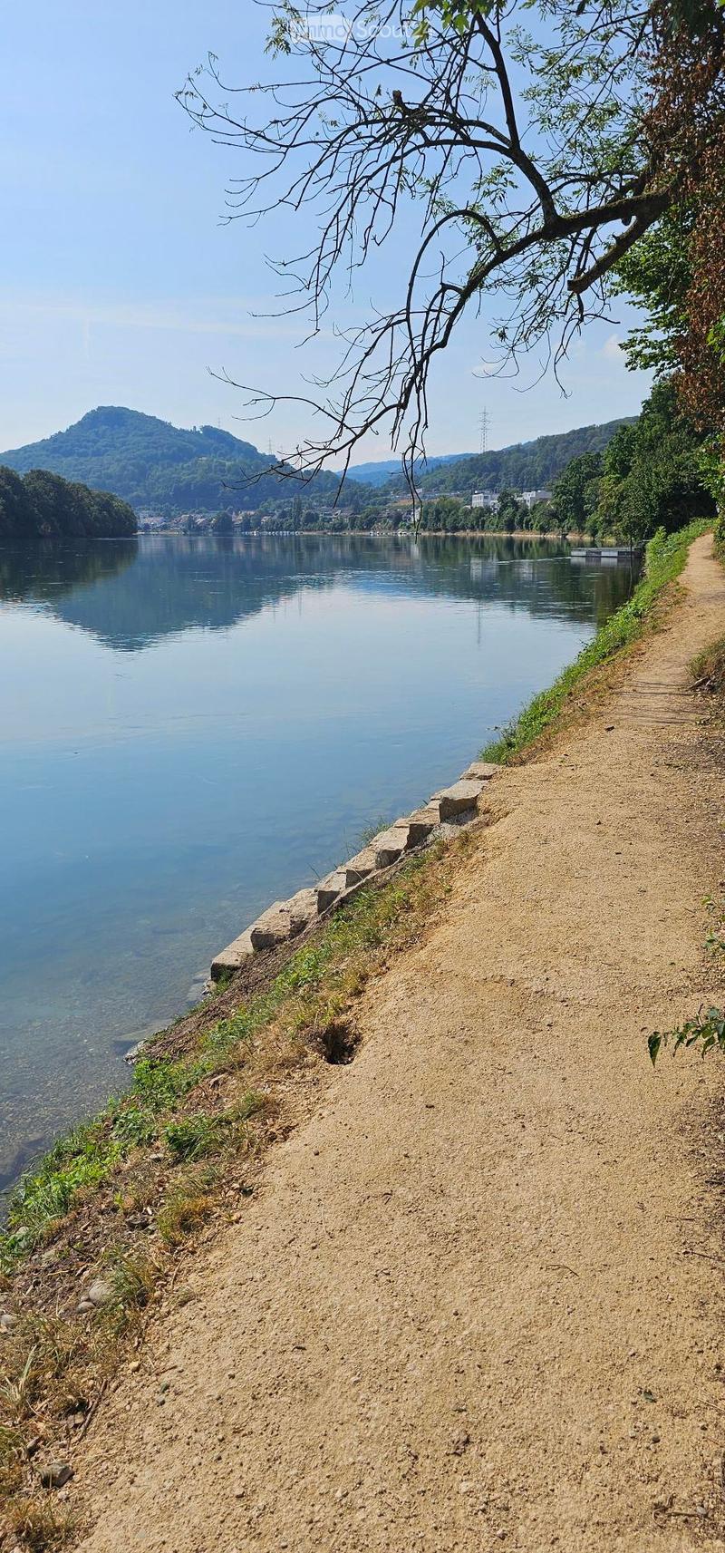 A serene riverside path with a gravel surface, bordered by grass and a stone edge, overlooking a calm river with distant hills and trees.