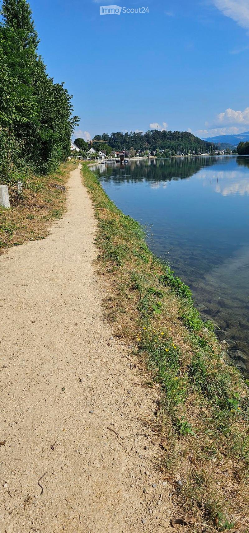 A gravel path runs alongside a calm river, with grassy banks and distant hills under a clear blue sky.