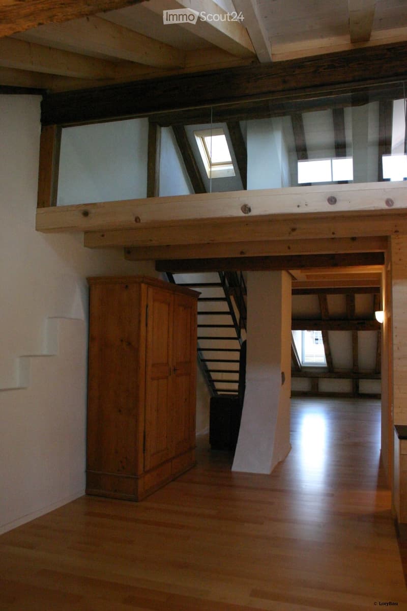 Empty room with wooden floor, exposed wooden beams, and a staircase leading to an upper level. There is a wooden cabinet on the left side and a small landing area with a window above.