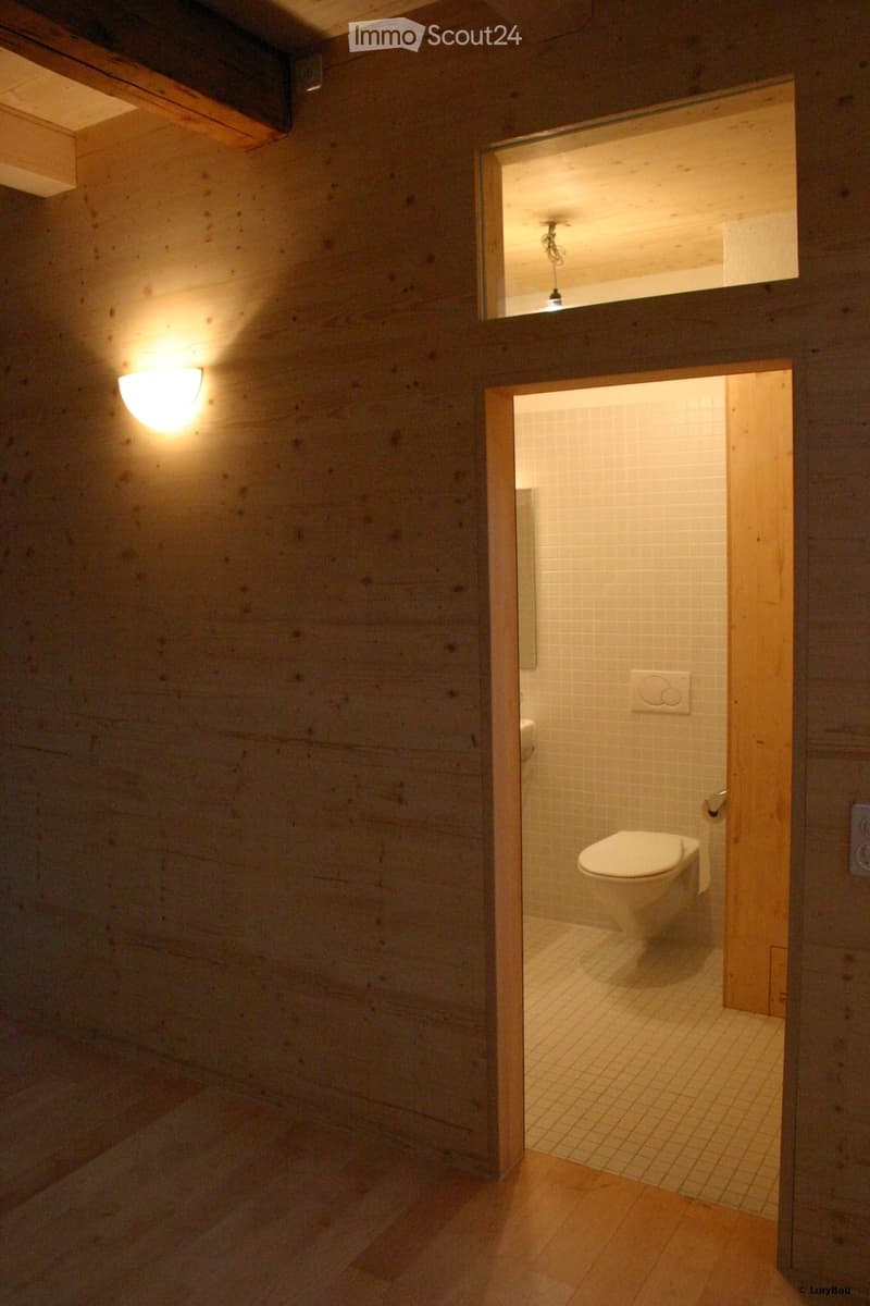 A small, modern bathroom with wooden walls and a white tiled floor. The toilet is visible, and the space is illuminated by warm lighting.
