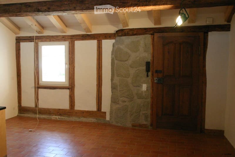 Empty room with exposed wooden beams, stone wall, wooden door, and a single window letting in natural light.