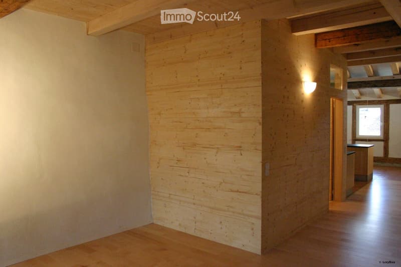 Empty hallway with wooden flooring and exposed wooden beams on the ceiling. The walls are painted in neutral tones, and there is a small light fixture mounted on one wall. The hallway leads to another room with a visible window and part of a kitchen area.
