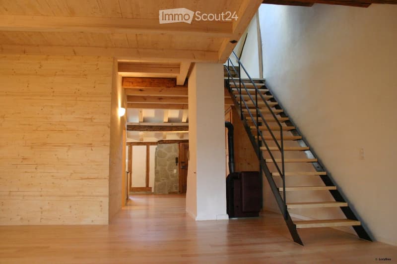 Spacious hallway with wooden flooring, exposed wooden beams, and a staircase leading upwards. Natural light filters through a window on the right side.