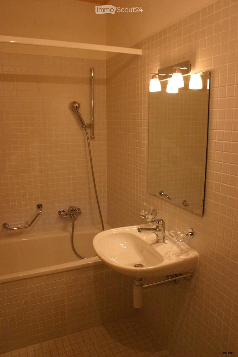 Bathroom with a white tiled wall and floor, a sink with a faucet, a bathtub with a handheld shower, a large mirror with built-in lighting, and a towel rack.