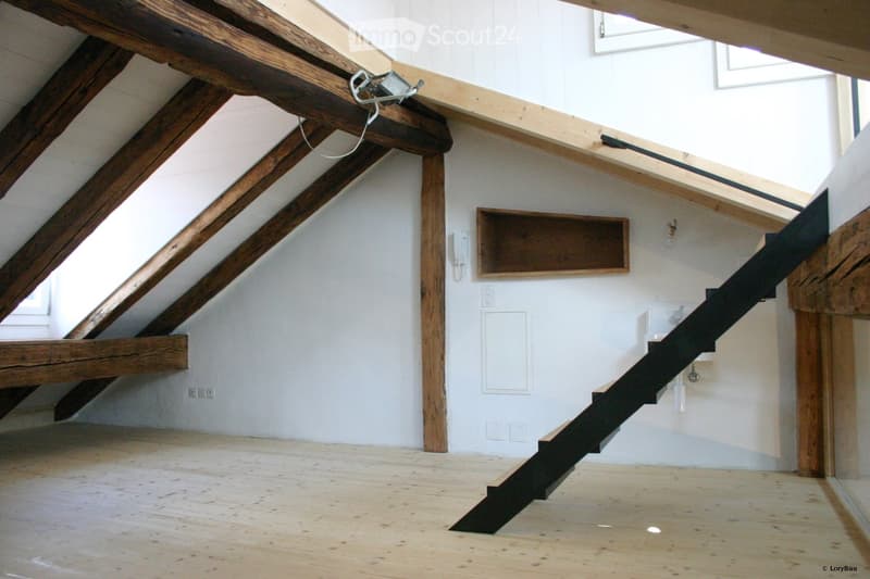 An attic space with exposed wooden beams, a wooden staircase leading up, and white walls. The floor is made of light-colored wood planks.