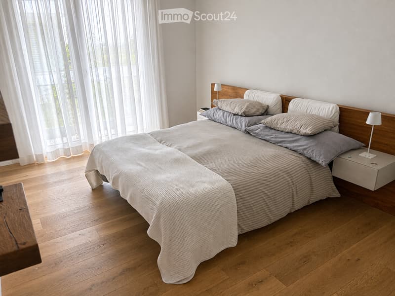 A modern bedroom with a large bed, wooden headboard, nightstand, and lamp. The room features light-colored bedding and wooden flooring.