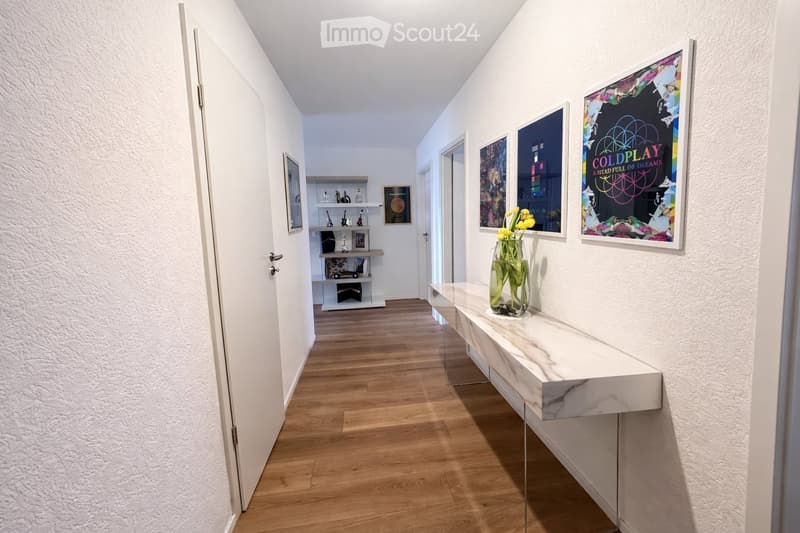 A modern hallway with wooden flooring, white walls, and a marble console table holding a vase of yellow flowers. Several framed artworks are displayed on the walls, and a small shelf unit is visible in the background.
