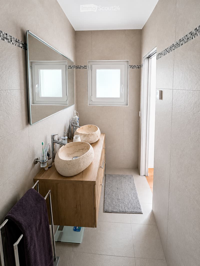 Modern bathroom with two stone sinks, wooden vanity, large mirror, and tiled walls.