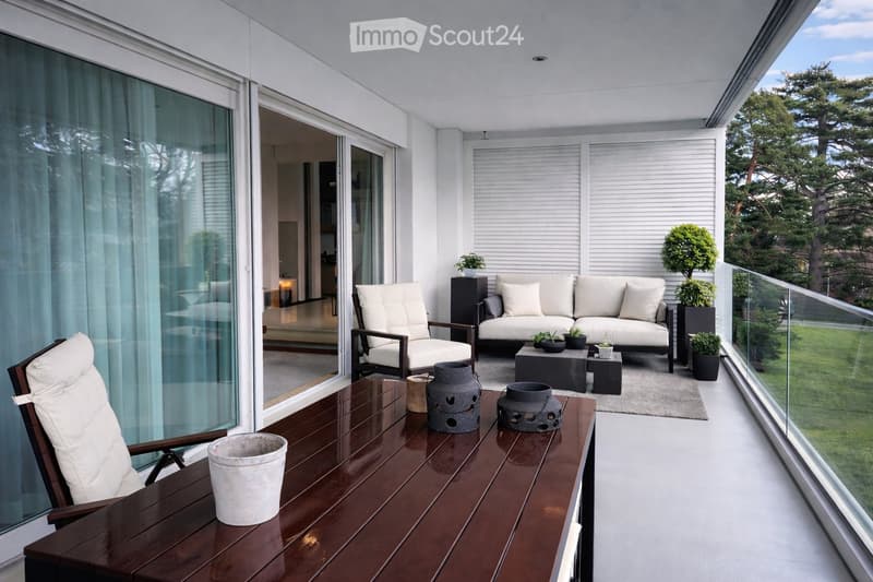 Modern balcony with white furniture, wooden table, potted plants, and a view of greenery.