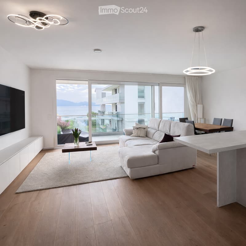 Modern living room with a large sectional sofa, coffee table, dining area, and a view of the sea through large windows and a balcony.