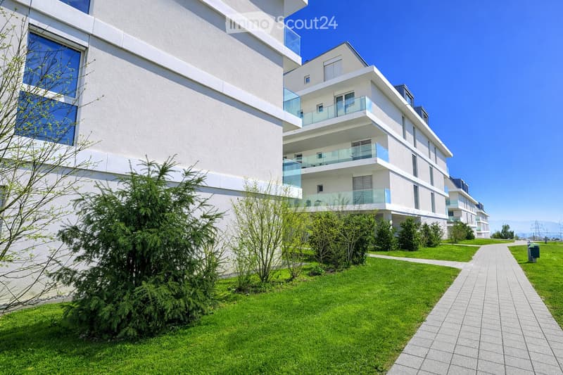 Modern multi-story residential building with white facade, large windows, and balconies. The surrounding area features a well-maintained lawn, small trees, and a paved walkway.