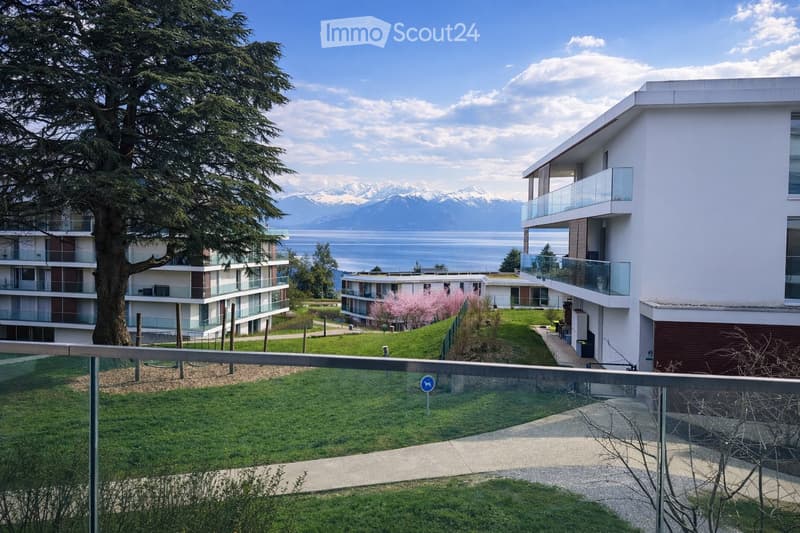 Modern residential complex with multiple buildings, balconies, and a scenic view of mountains and a lake in the background.