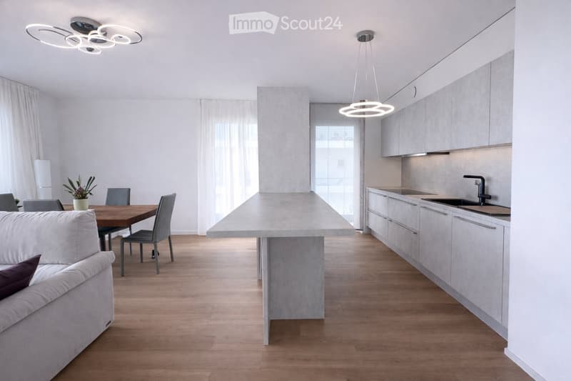 Open-plan living and dining area with a modern kitchen. Features a white sofa, wooden dining table with chairs, a central kitchen island, and sleek kitchen cabinets.