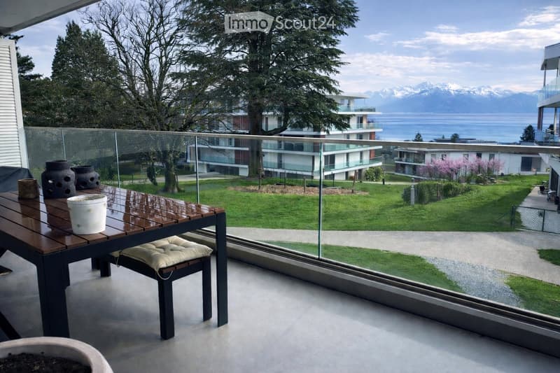Balcony with a wooden table, a stool, and vases, overlooking a landscaped garden and distant mountains.