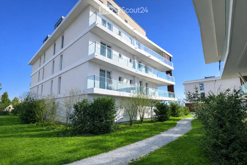 Modern multi-story residential building with white facade, large windows, and glass balconies. The building is surrounded by a well-maintained lawn and gravel pathway.