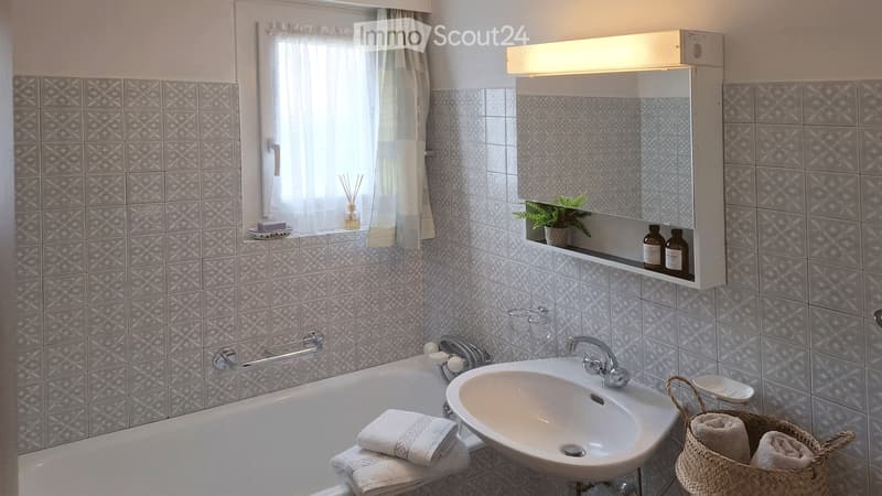 Bathroom with grey tiled walls, a bathtub, a sink, a mirror cabinet, and a window with a curtain. Towels are placed on the edge of the bathtub, and a basket with towels is on the floor.