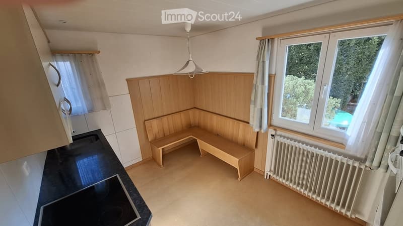 A compact, well-lit room with a wooden bench seating area, a black kitchen counter with an induction stove, and large windows with curtains. The walls are paneled in light wood, and a radiator is visible beneath the windows.