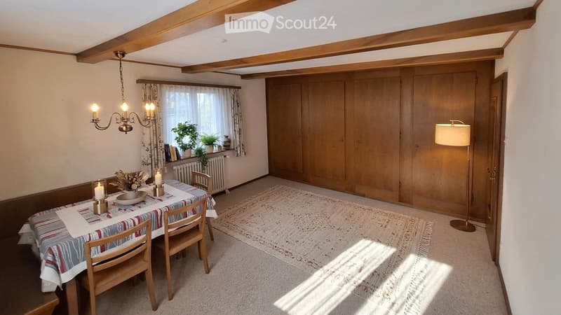 Cozy dining area with wooden dining table, chairs, chandelier, floor lamp, and a large window with curtains. Wooden cabinetry and a rug complete the space.