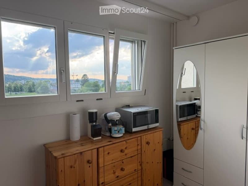 A small, well-lit kitchen area with wooden cabinetry, a microwave, coffee maker, and a refrigerator with a built-in microwave. The room has large windows offering a scenic view of the landscape.