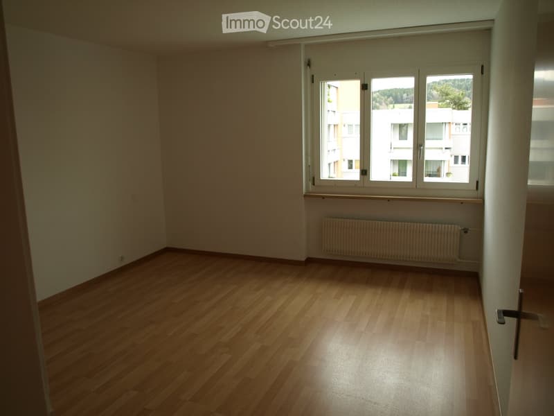 Empty room with wooden flooring, large windows, radiator, and a door handle visible.