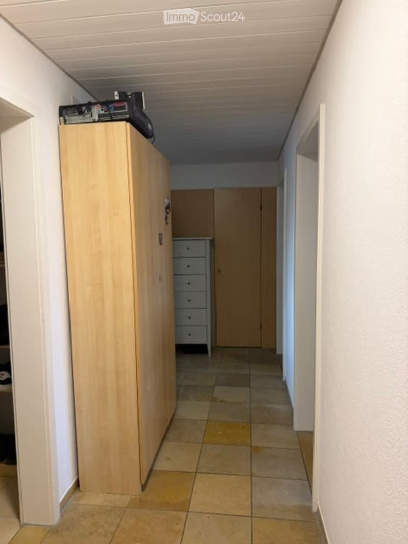 A narrow hallway with tiled flooring, white walls, and a wooden wardrobe on the left side. A small cabinet is visible further down the hallway.