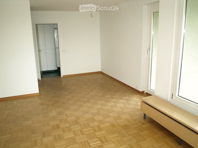 Empty room with wooden parquet flooring, white walls, and a small bench near a window. The room has a doorway leading to another space with cabinets.