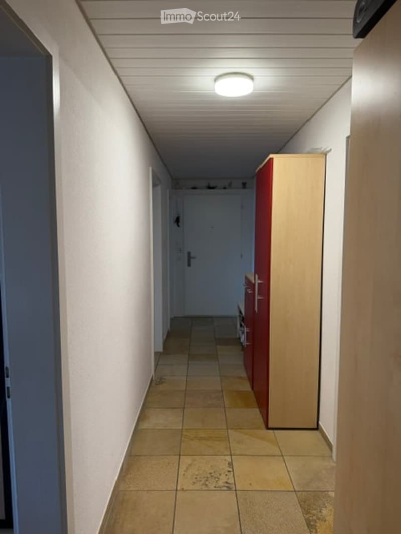 A narrow hallway with tiled flooring, white walls, and a ceiling light. There are doors on both sides and a small cabinet on the right.